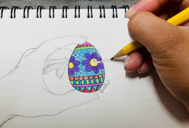 A photograph of a person drawing an egg. The person is using a pencil to draw a colorful egg being held by a person's hand.
